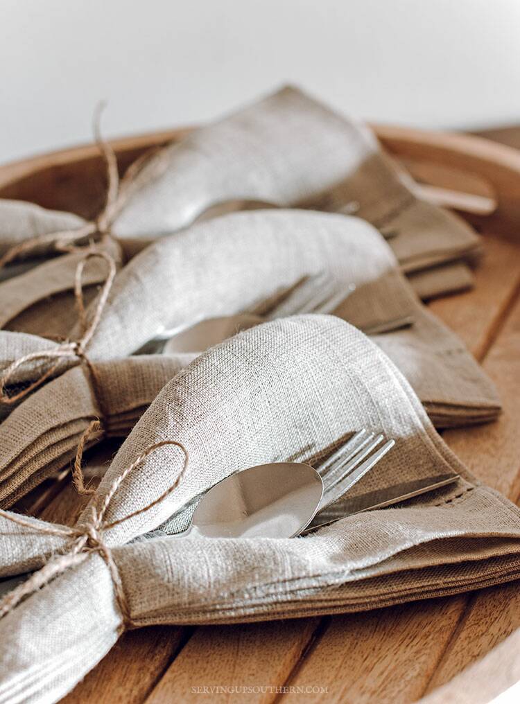 Three sets of cutlery wrapped in cloth linen napkins on a wooden tray.