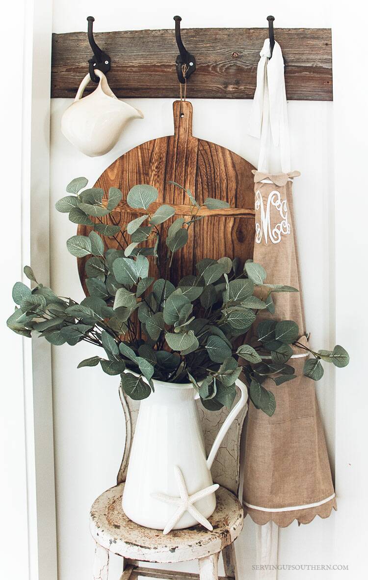 White metal pitcher full of eucalyptus sitting on a vintage metal stool with a large round breadboard hanging in the background.