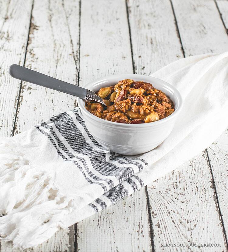 A bowl of calico beans sitting on a pretty dishcloth on a white weathered wooden board