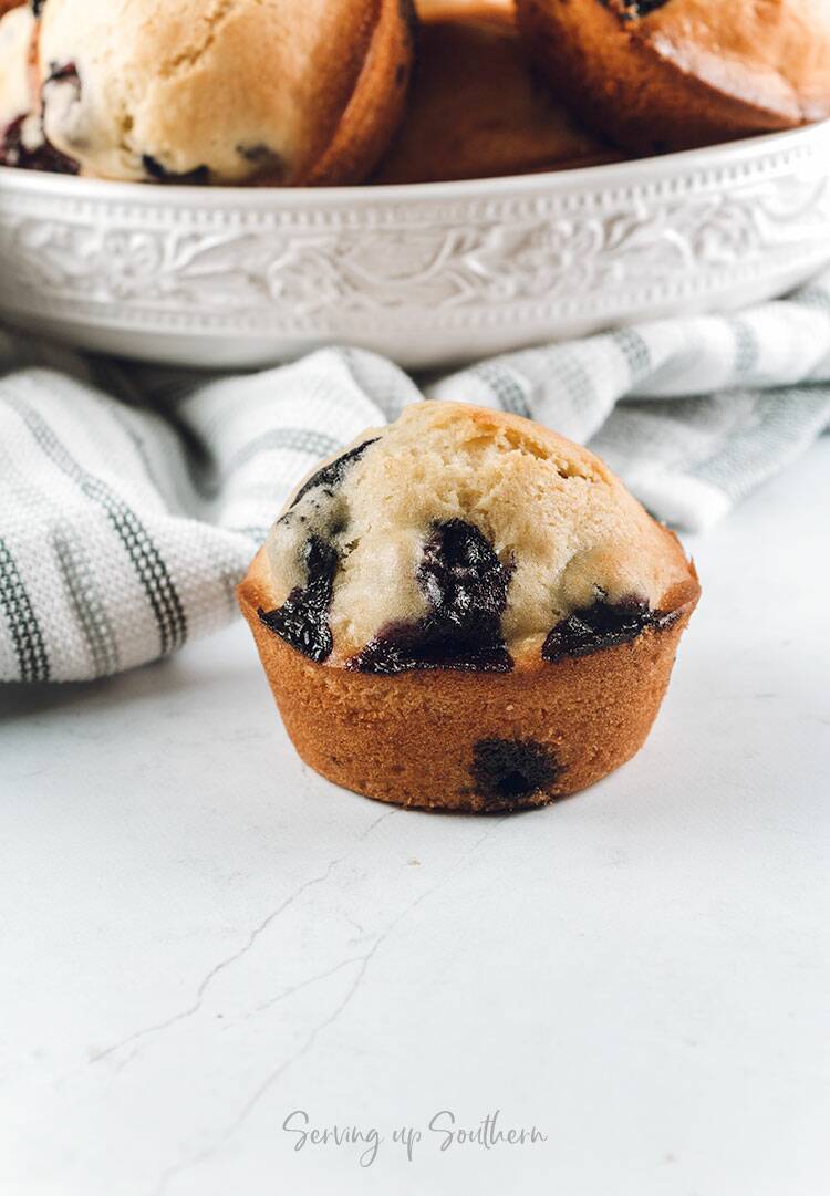 A blueberry muffin on a marble surface with a bowl of muffins in the background.