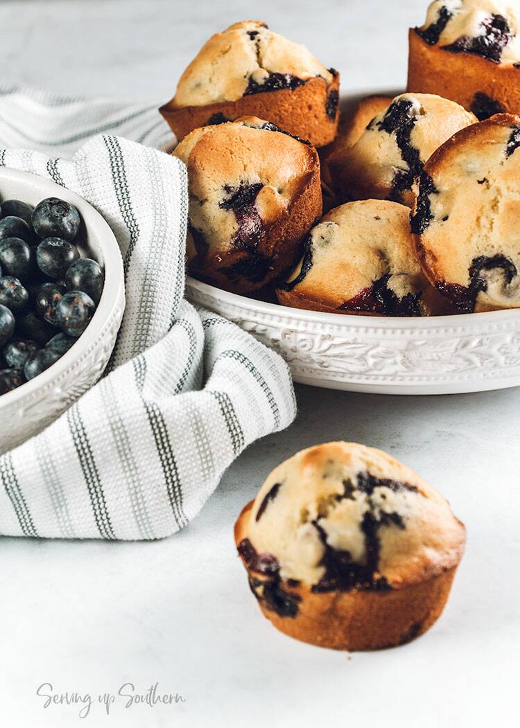 Blueberry muffin on marble surface with a bowl of fresh blueberries and baked muffins in the background.