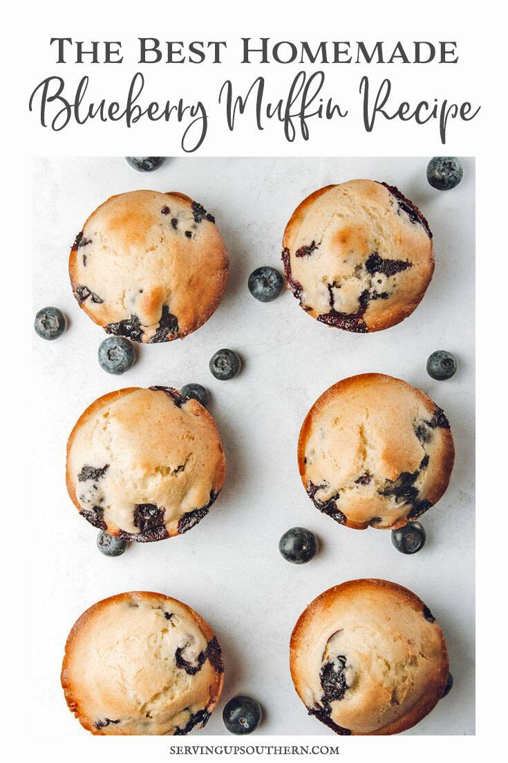 Six blueberry muffins on a marble top with fresh blueberries scattered around them.