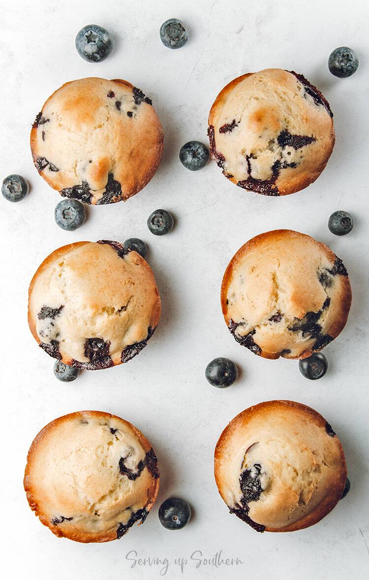 Six blueberry muffins on a marble top with fresh blueberries scattered around them.