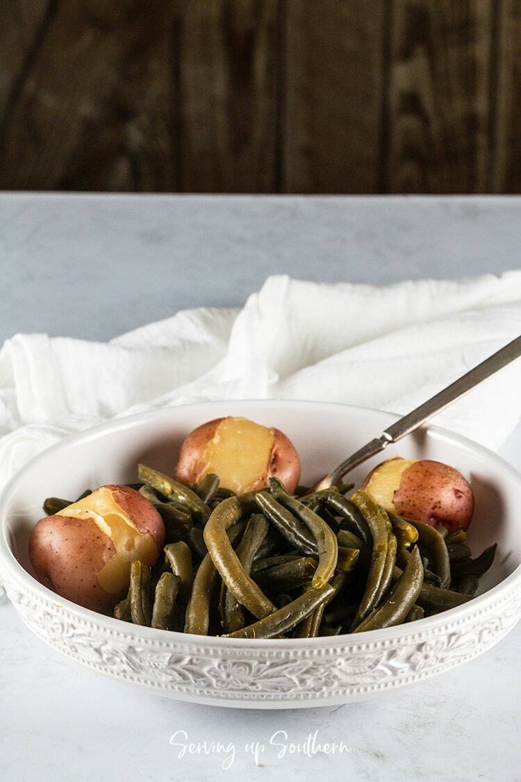 Picture of a white bowl filled with southern style green beans and new potatoes on a marble surface.