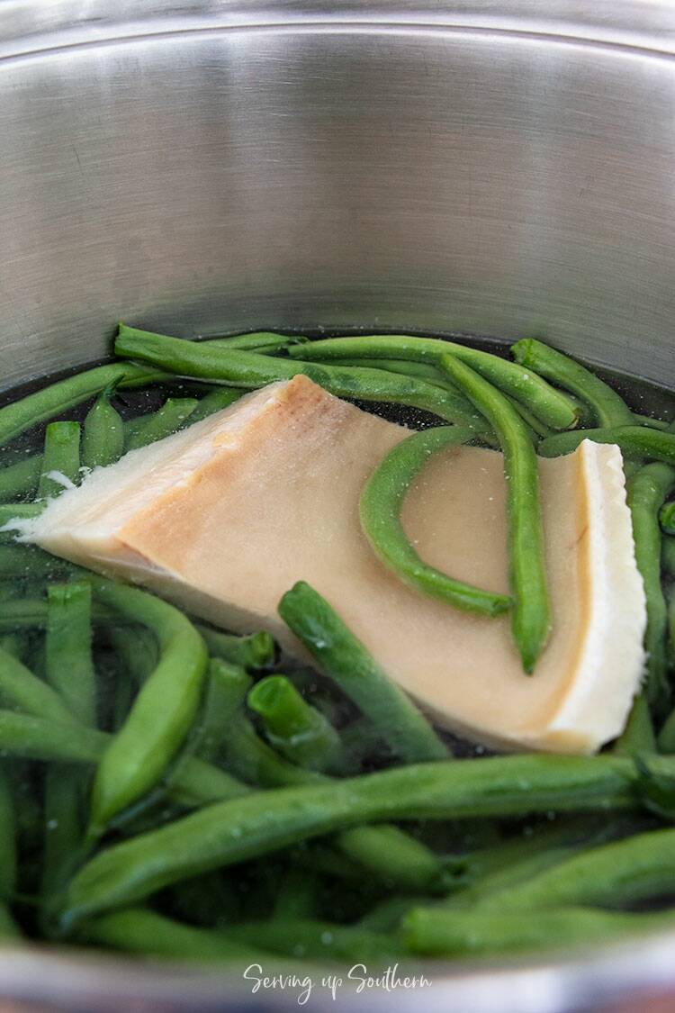 Freshly snapped green beans and a slab of fatback in a stainless steel pot with water.