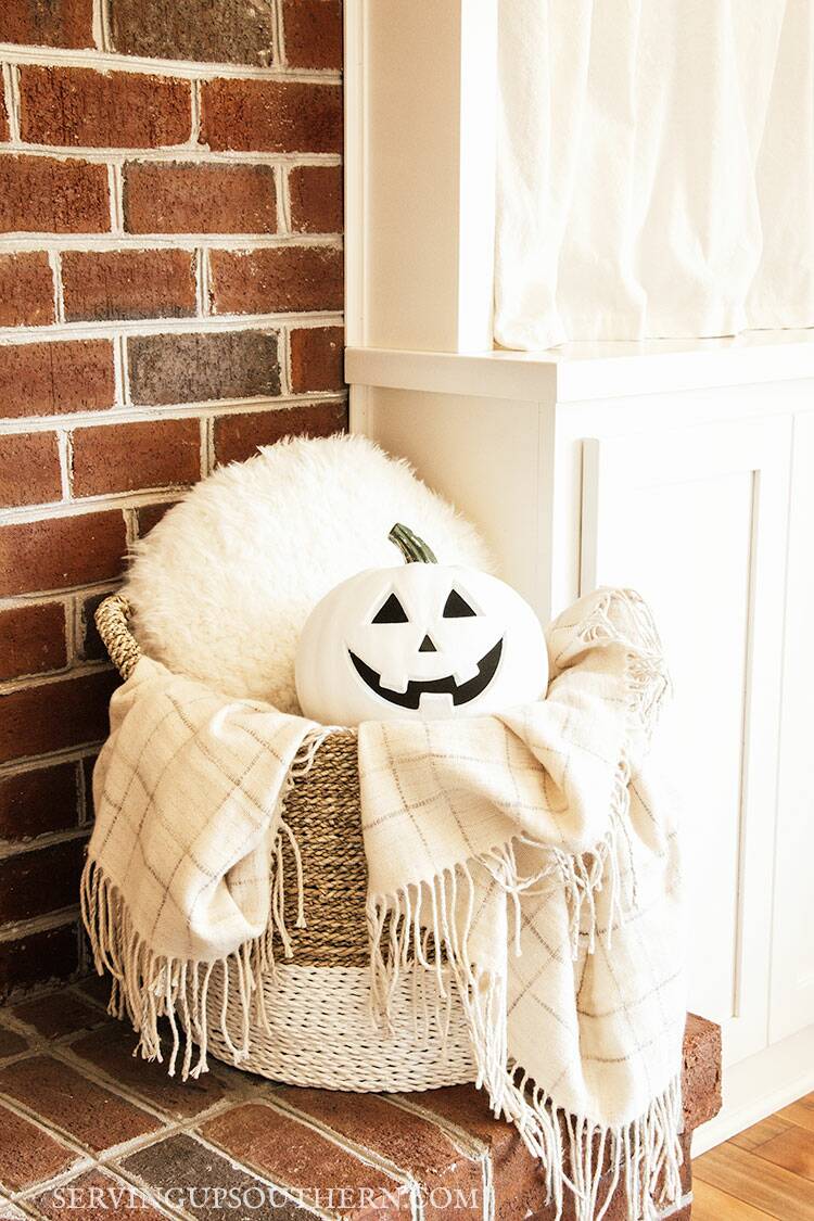 White jack o' lantern nestled in a basket with a pillow and throw sitting on a hearth.