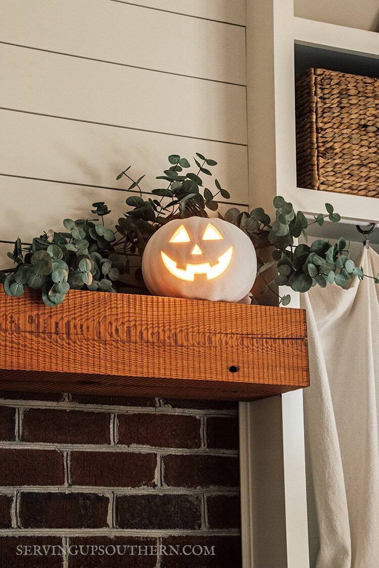 White painted Jack O' Lantern lit up and sitting on a fireplace mantel with eucalyptus.