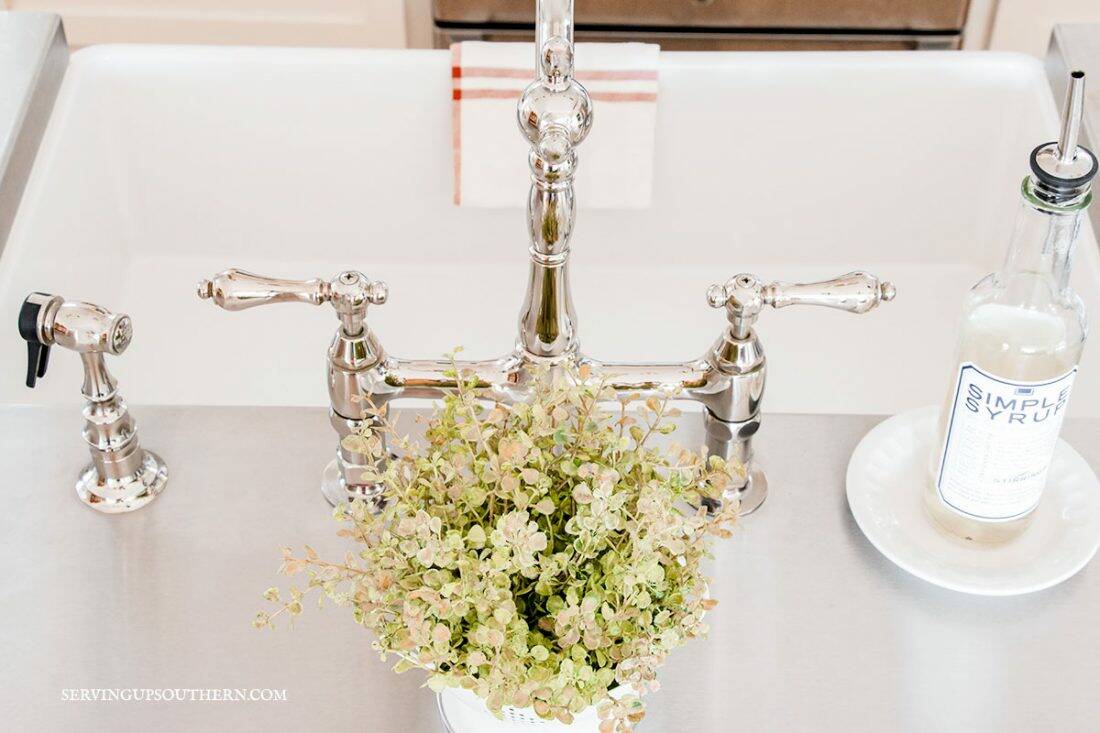 Close-up picture of a farmhouse sink with a beautiful bridge faucet surrounded by stainless steel countertops and a small green plant.