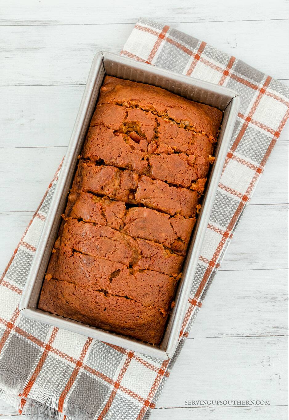 A loaf pan with sliced pumpkin bread sitting on a white wooden board with a gray and rust colored dish towel.