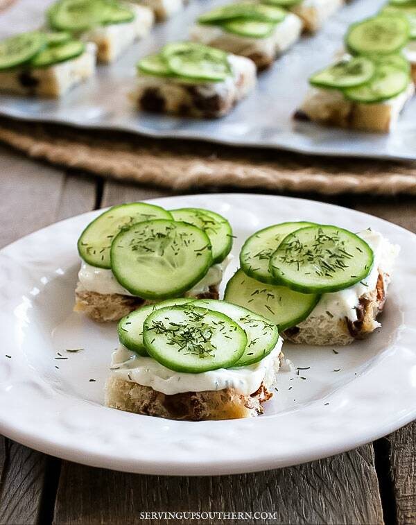 Fancy cucumber tea sandwiches on a white plate on a wooden board.