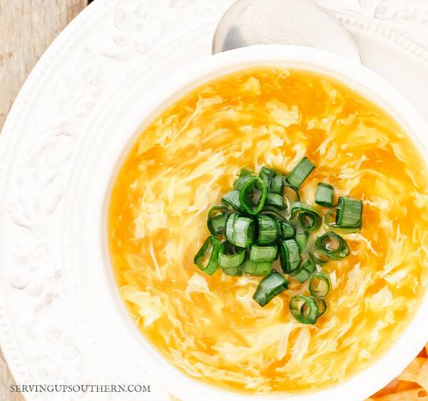 Egg drop soup topped with sliced green onions in a white bowl on a wooden board.