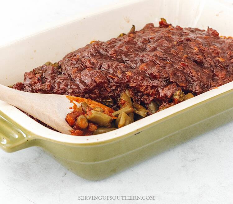 A green vintage baking dish of barbecued green beans with a wooden spoon sitting on a white marble surface.