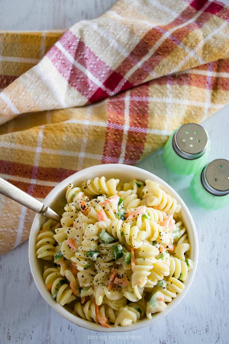 Amish Macaroni Salad in a white bow with jade salt & pepper shakers and a napkin on a white rustic board.