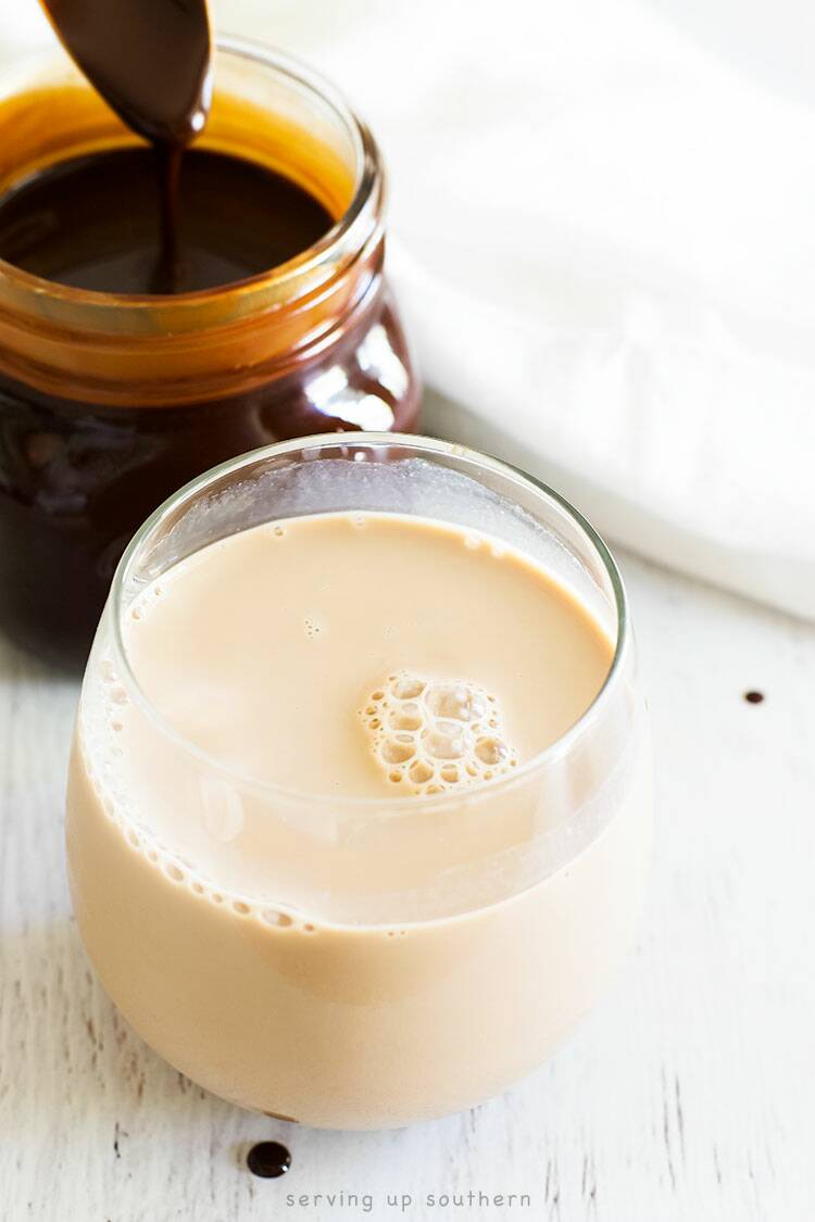 A glass of chocolate milk sitting next to a jar of homemade chocolate syrup on a white wood board.