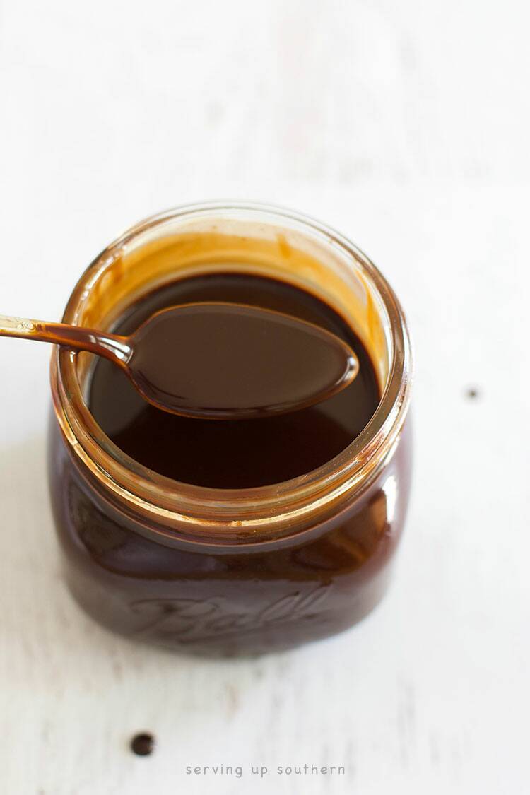 A jar of homemade chocolate syrup in a half-pint mason jar with a spoon on a white wood board.