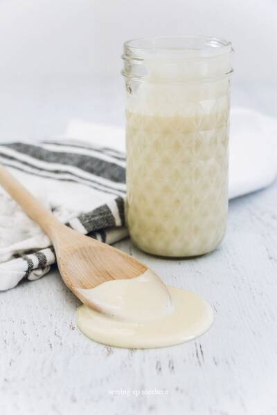 A jar of homemade sweetened condensed milk and a wooden spoon with a tea towel on a white wood board.