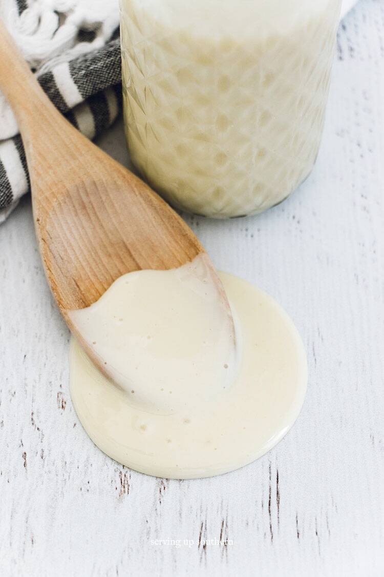 A jar of homemade sweetened condensed milk and a wooden spoon with a tea towel on a white wood board.