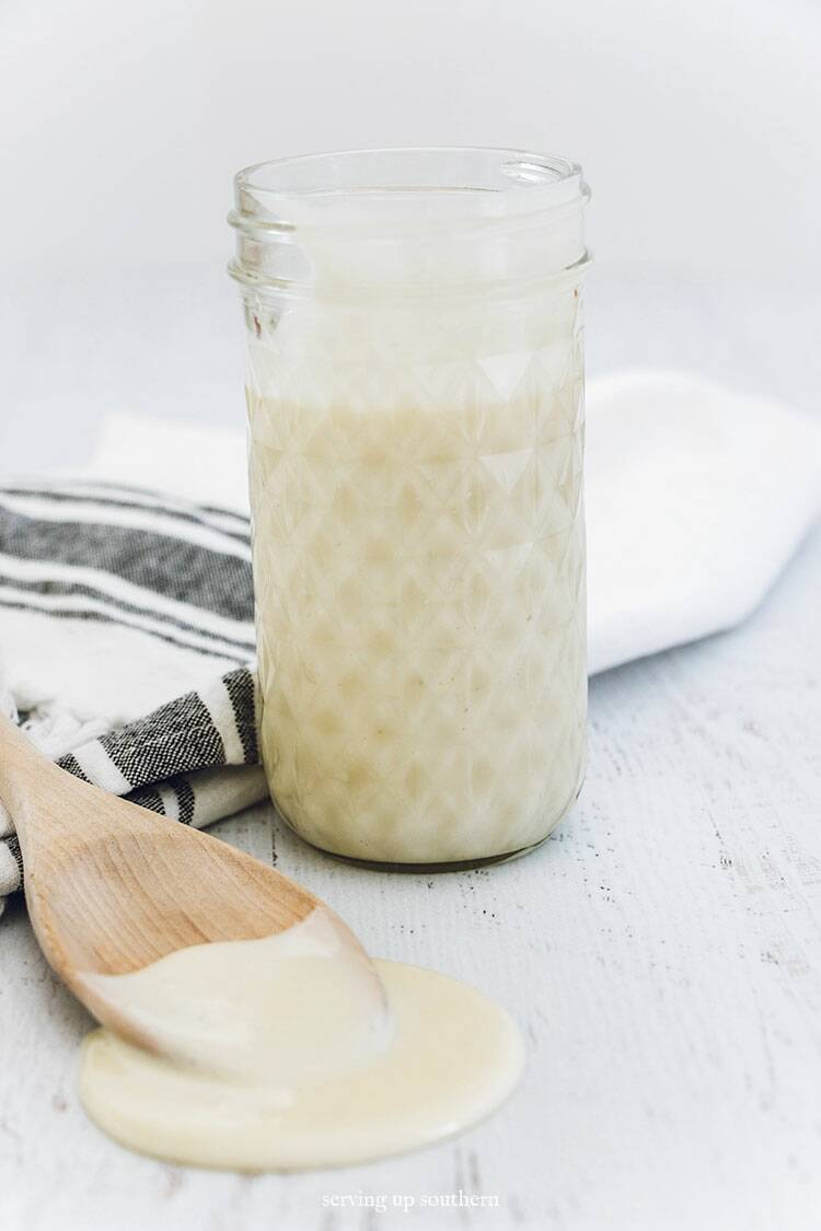 A jar of homemade sweetened condensed milk and a wooden spoon with a tea towel on a white wood board.