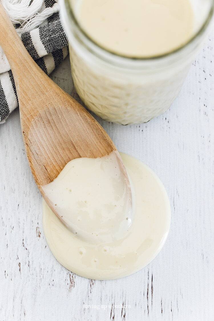 A jar of homemade sweetened condensed milk and a wooden spoon with a tea towel on a white wood board.