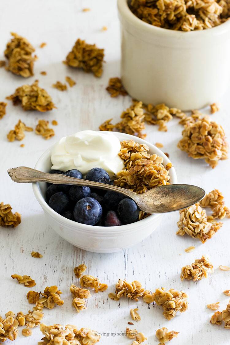 A white bowl filled with homemade granola, greek yogurt, and blueberries sitting on a white wooden board.