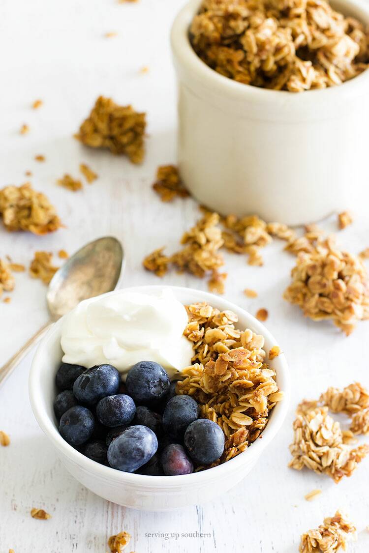 Picture of a bowl on homemade granola in a white bowl with blueberries, and greek yogurt on a white wooden board.