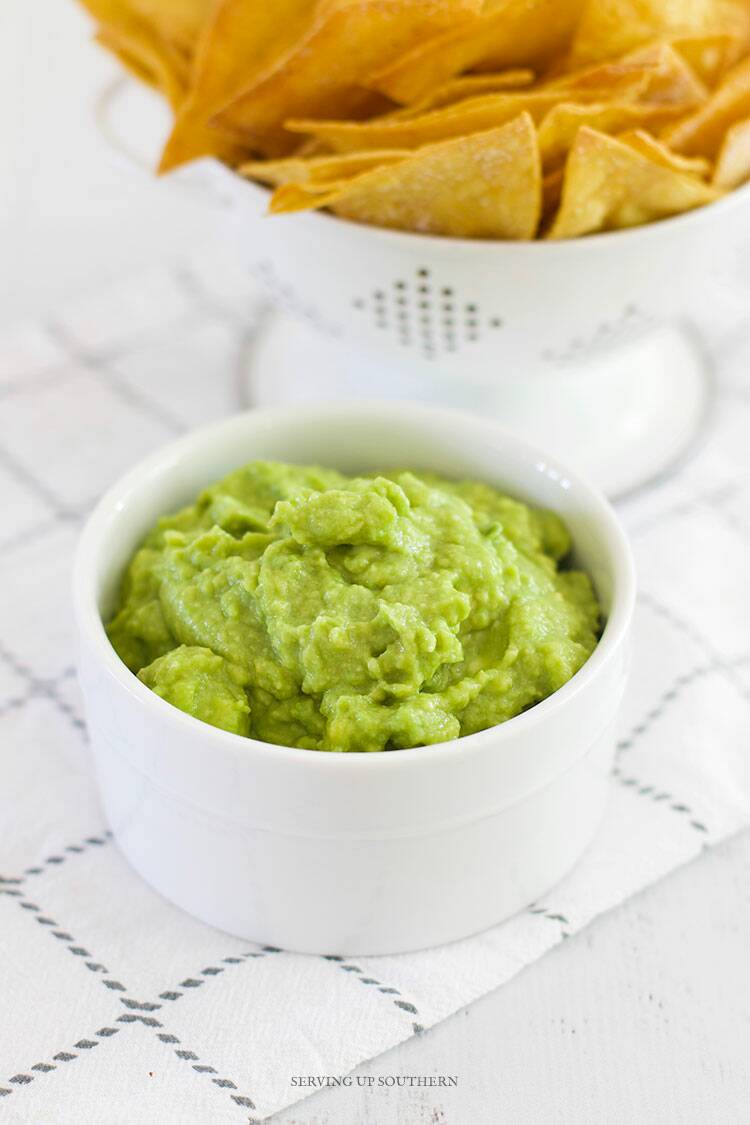 A small white bowl of homemade guacamole sitting on a white wood board with chips.