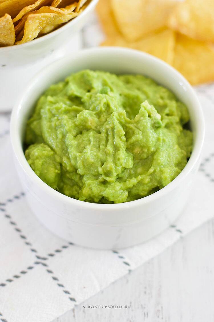 A close up shot of guacamole dip in a white bowl with chips on the side.
