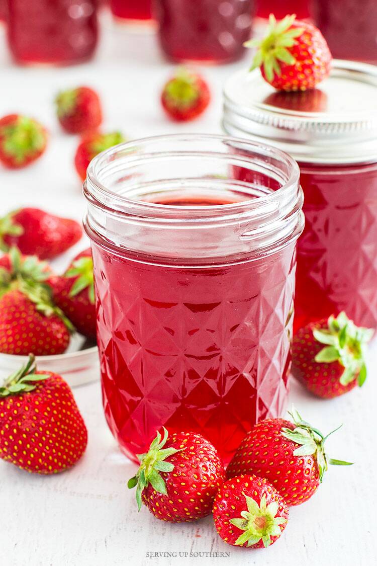 Homemade strawberry syrup in a quilted jelly jar with fresh strawberries scattered about.