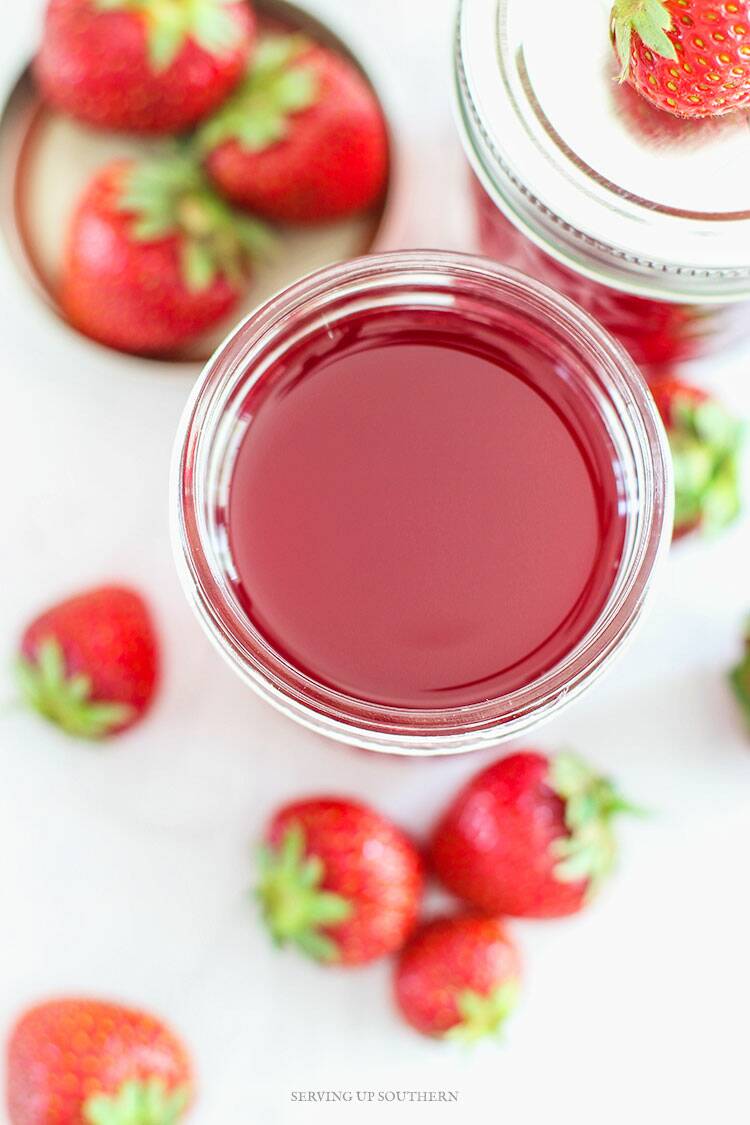 Jelly jar filled with homemade strawberry simple syrup with fresh strawberries scattered about.