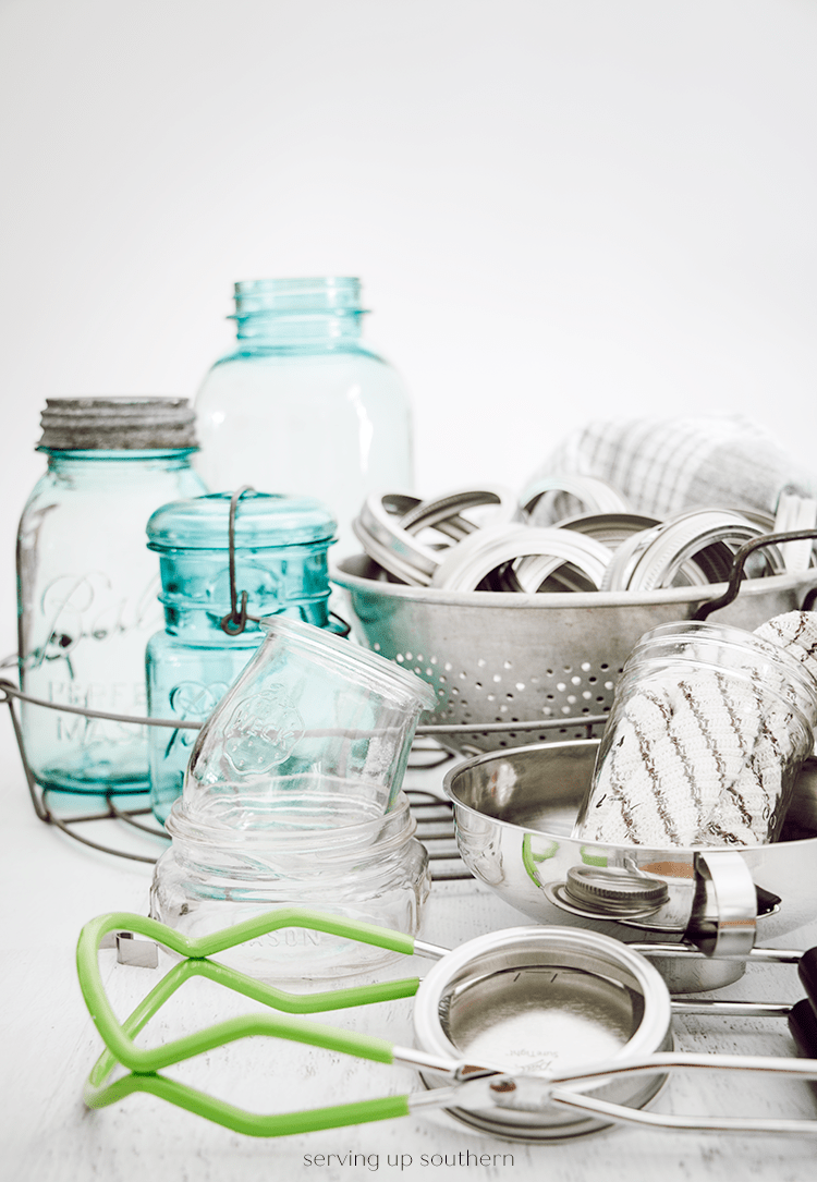 An image of canning supplies including vintage blue ball jars, clear jars, rings, colander, jar lifter, funnel,  dish towel, and a dish cloth.