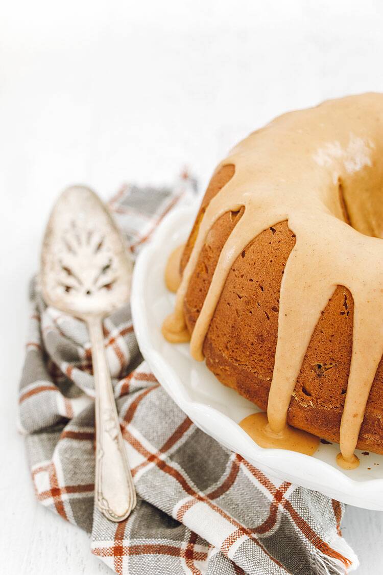 Pumpkin Bundt Cake drizzled with pumpkin spice glaze on a white cake plate with a sliver cake server and dish towel.