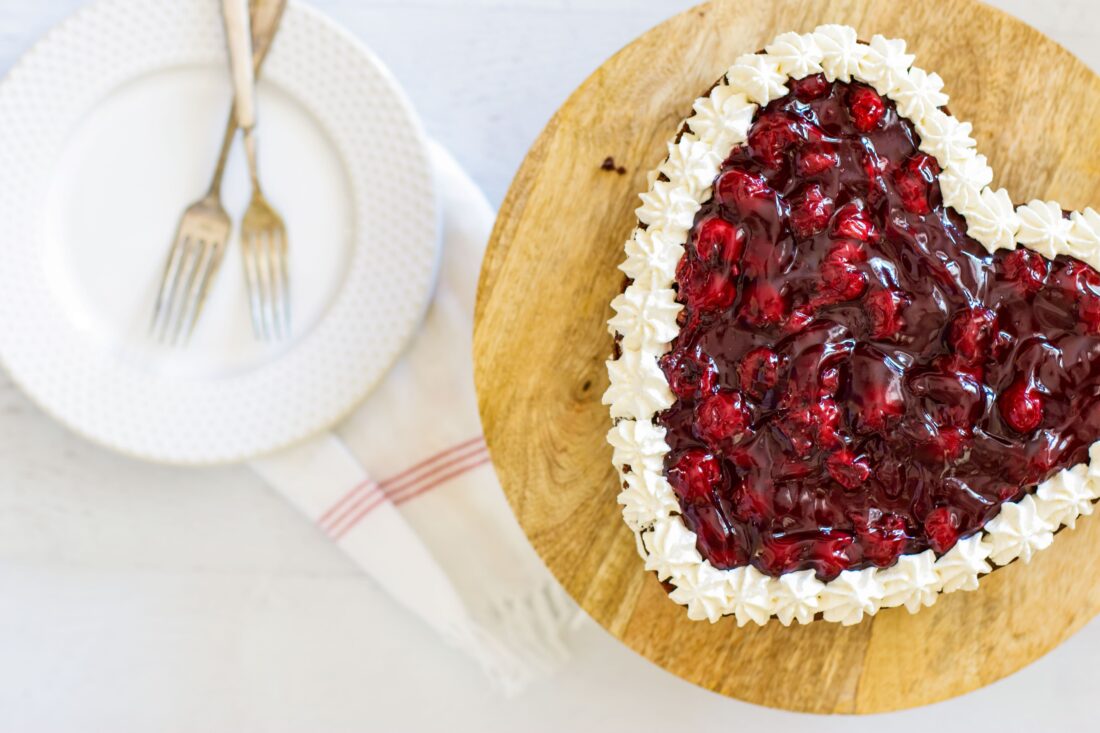 Heart shaped chocolate cake topped with cherry pie filing on a wooded cake stand with plates and forks on the side.