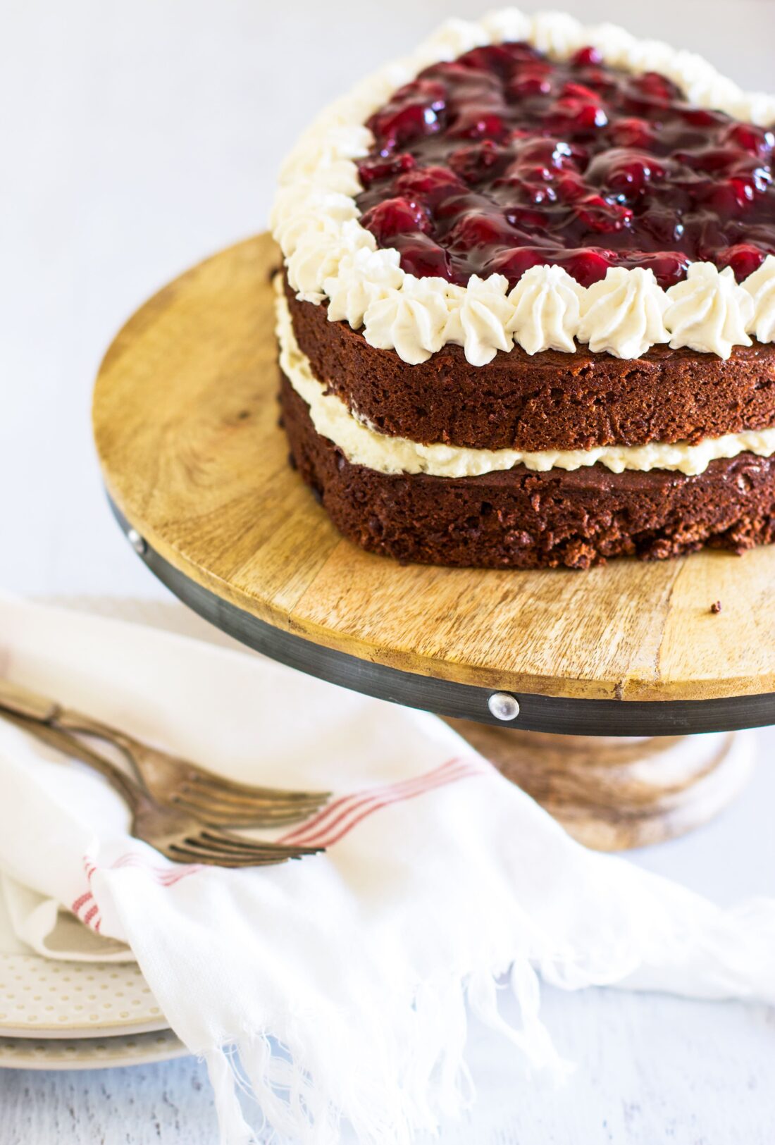 Heart shaped chocolate cake topped with cherry pie filing on a wooded cake stand with plates and forks on the side.