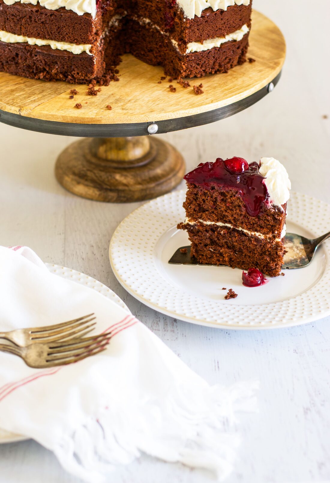 Slice of chocolate cake topped with cherry pie filling on a white plate in front of cake on pedestal.