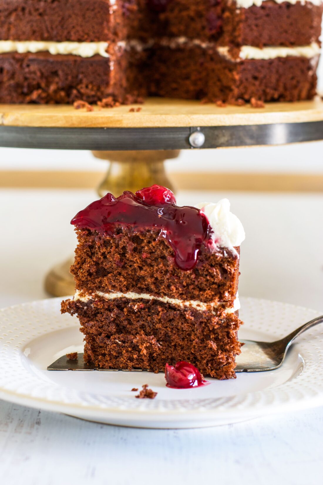 Slice of chocolate cake topped with cherry pie filling on a white plate in front of cake on pedestal.