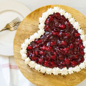Heart shaped chocolate cake with cream filling and topped with cherry pie filling on a wooden cake stand.