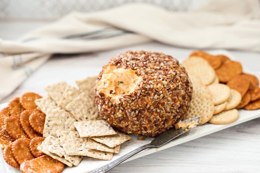 Cheese ball on a white plate with crackers on a white board.