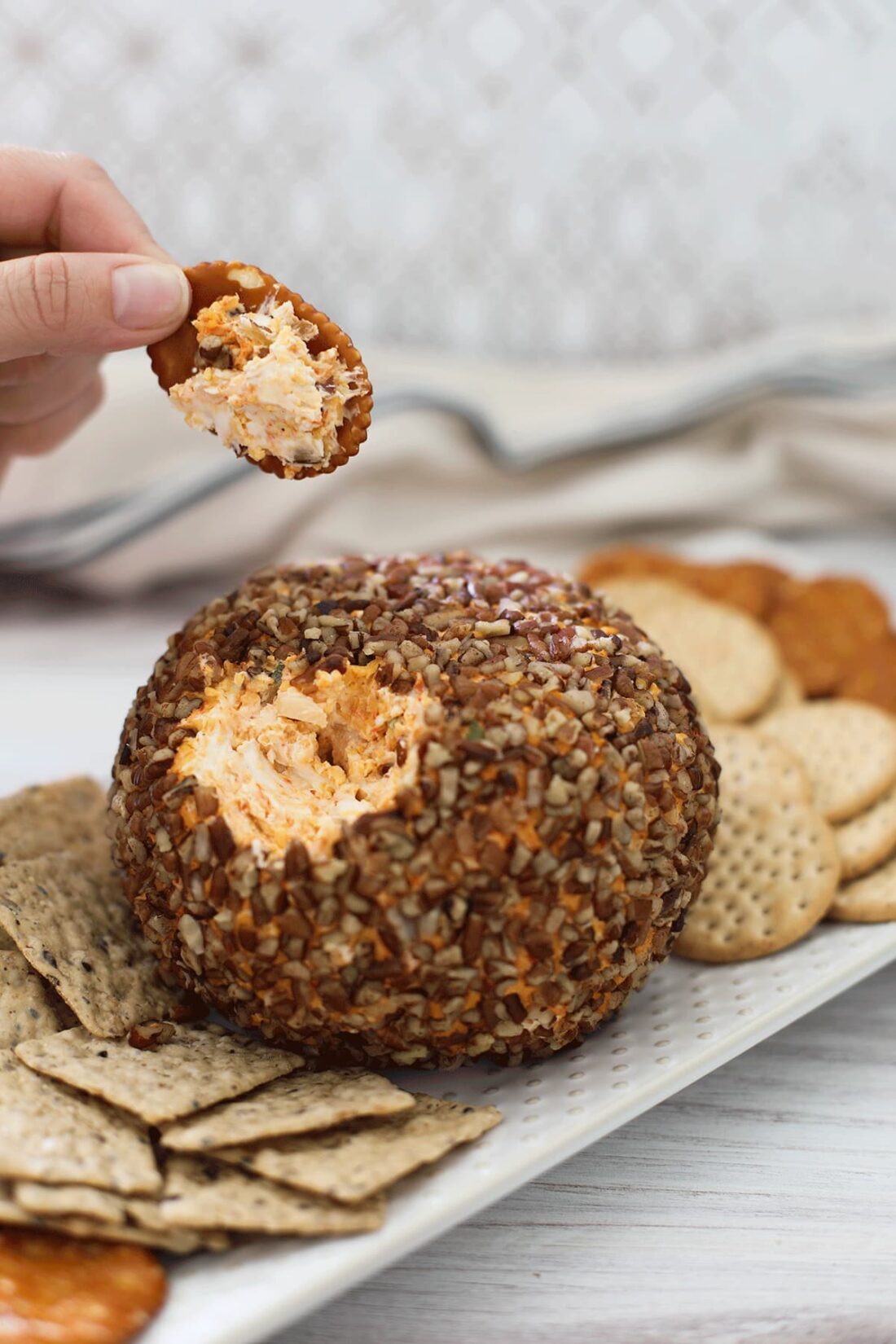 Cheese ball on a white plate with crackers on a white board with someone holding a cracker spread with cheese.