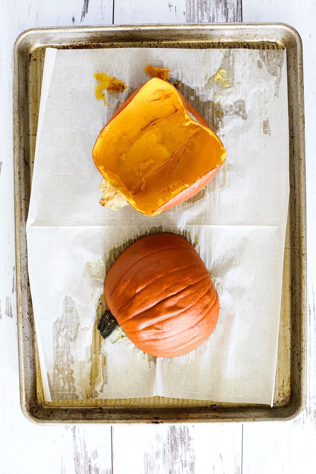 Baked sugar pumpkin halves on a parchment paper-lined sheet pan. One halve has the pulp scooped out.